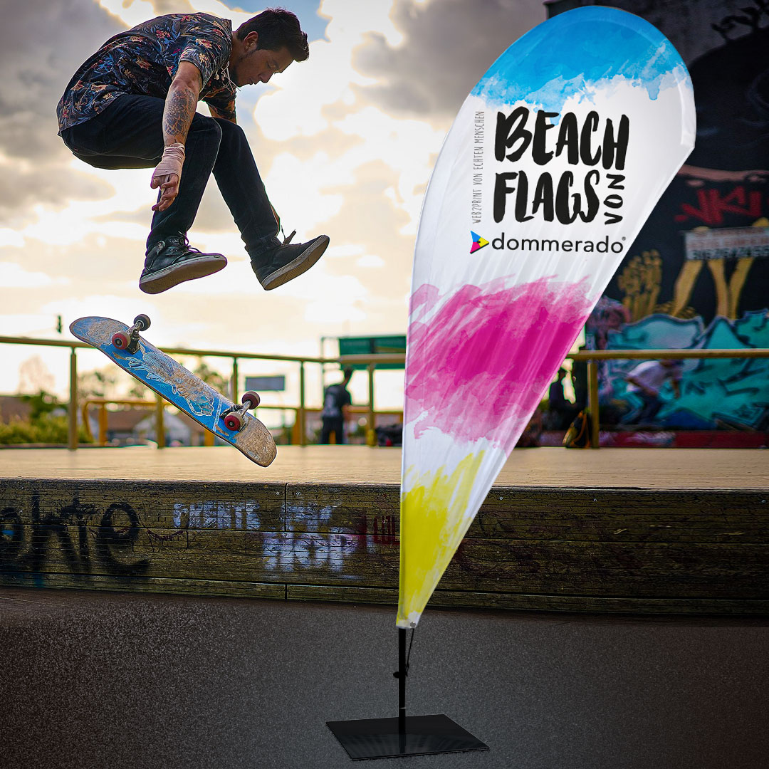 Beachflag in Tropfenform mit farbigem Motiv, auf Bodenplatte vor einer Halfpipe im Außenbereich, im Hintergrund springt eine Person mit Skateboard
