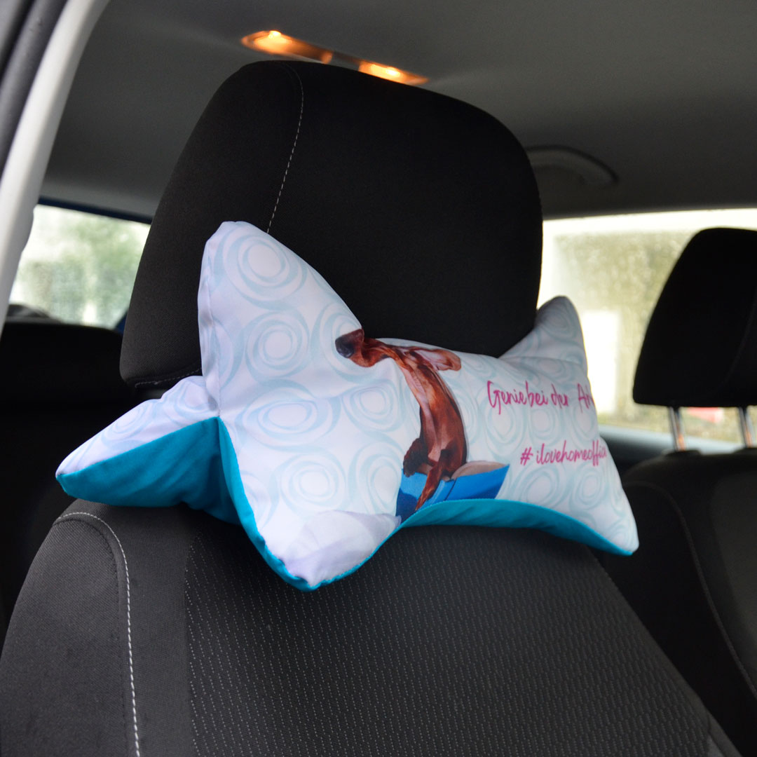 Ein bedrucktes Nackenkissen mit Dackel-Motiv und Spruch ist an einer Auto-Kopfstütze befestigt.