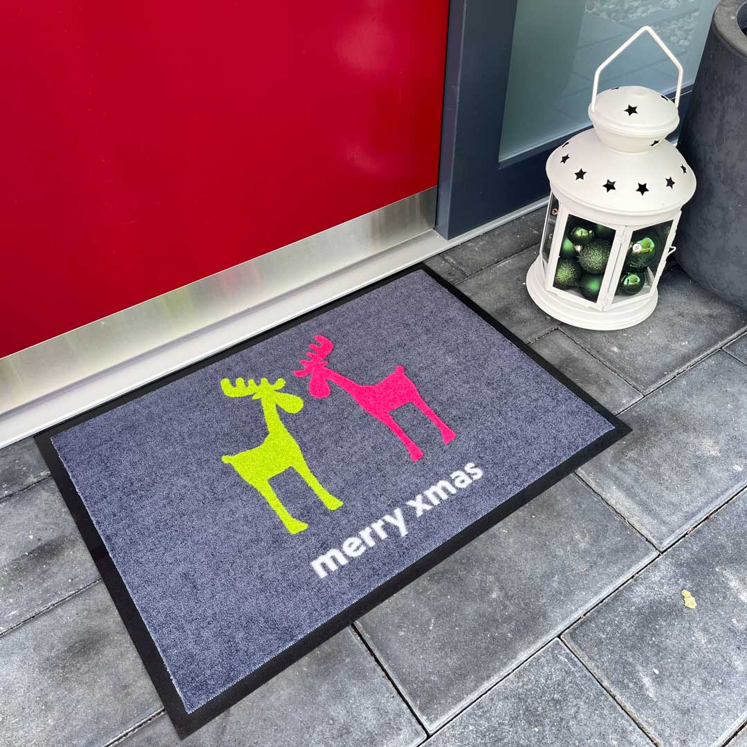 Eine graue Fußmatte mit einem bunten Weihnachtsmotiv (zwei Rentiere in Neongelb und Pink) und der weißen Aufschrift "merry xmas", die vor einer roten Tür liegt.